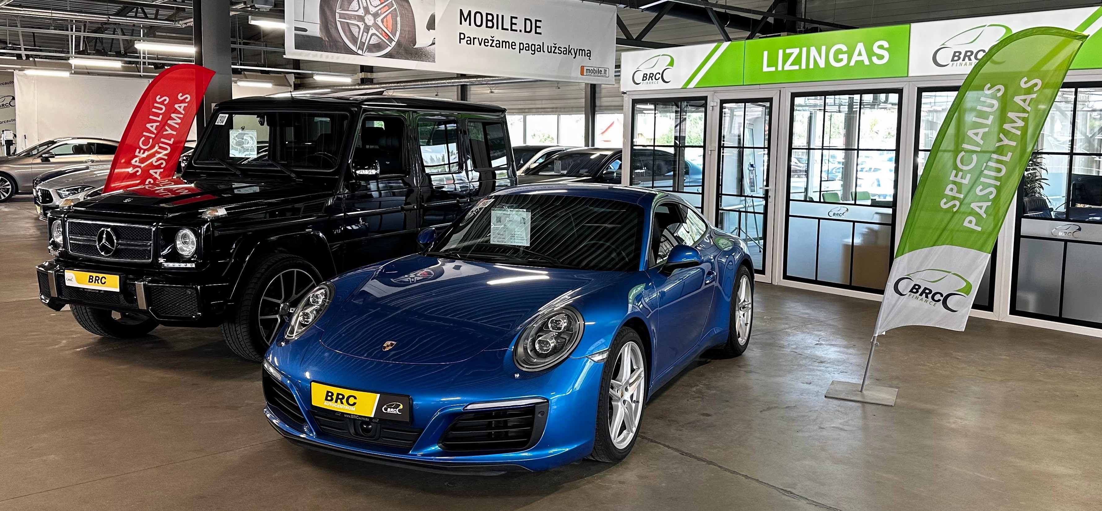 Auto salona ekspozīcija ar melnu Mercedes-Benz G-Class apvidus auto un zilu Porsche sporta automašīnu, kas novietotas blakus. Apkārt redzamas BRC izkārtnes, karogi ar uzrakstu ‘Īpašais piedāvājums’ un biroja stikla starpsienu zona.